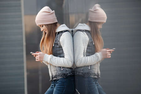Cute girl with blue eyes, long, straight, light brown hair, beautiful smile, dressed in a knit pullover, gray-blue color, wearing a sports cap pink, light makeup, spends time alone in the city, posing against the backdrop of the gray wall, spring portraitの写真素材