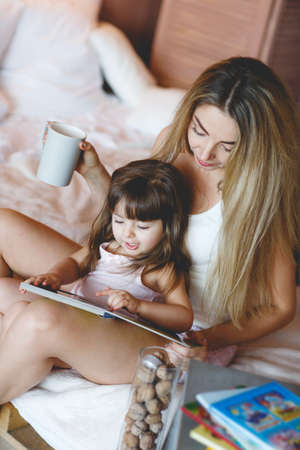 Young woman with long blonde straight hair and brown eyes and her daughter, girl-a brunette with brown eyes, 3 years old, lying in a white bed in the bedroomの写真素材