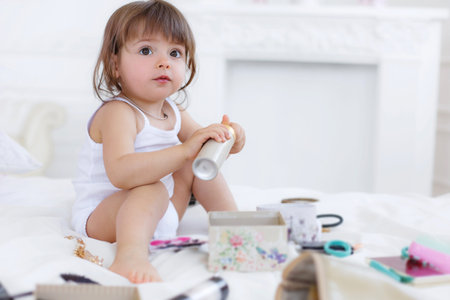 Happy little girl 3 years old, brunette with long hair and straight bangs, a nice smile and brown eyes sitting on a white bed in the bedroom, cross-legged amid the pile of white pillows with a huge bar of chocolate in hand, next to a Teddy bearの写真素材