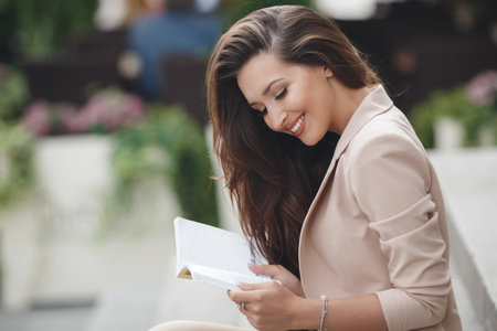Young beautiful woman brunette with long straight hair, long black eyelashes, and gray eyes, nice smile, straight white teeth and pink lipstick, wearing a pantsuit beige, wearing jewelry, reading a book, sitting on the steps outdoors in the cityの写真素材