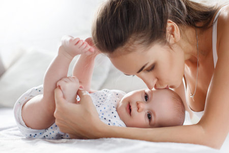 Beautiful young woman playing with her newborn son in the bedroom on the bed, a boy with a short,...