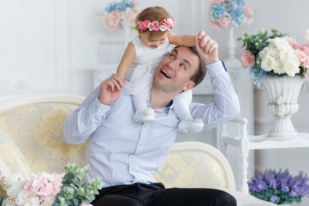 A young handsome man, a brunette with short hair, dressed in a white shirt and black pants, spends time at home with his little daughter, a girl with red hair, dressed in a white dress with a wreath on her headの写真素材