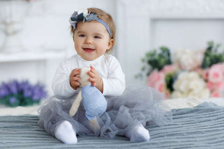 Little girl with brown eyes and red hair, but the head is wearing a gray-blue wreath with white and gray flowers, dressed in a white t-shirt and transparent gray-blue skirt, spends time alone, playing on a large bed in the bedroomの写真素材