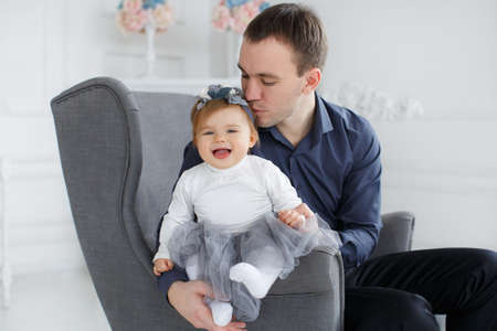 A young handsome man, a brunette with short hair, wearing a blue shirt and black pants, spends time at home sitting on the couch with his little daughter, a girl with red hair, dressed in a white dress with a wreath on her headの写真素材