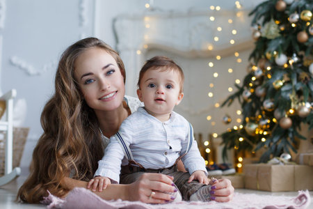 Happy family mother and little son near christmas tree on Christmas Eve at home. A woman and a little boy are resting in the white room near the Christmas tree. Family at home. A young mother playing with her son waiting for the New Yearの写真素材