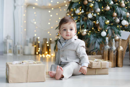 Little boy with huge brown eyes, blond short hair holding a toy for Christmas tree gold color, wearing a light brown trousers and white striped shirt, sitting on the floor next to a white Christmas tree and boxes with giftsの写真素材