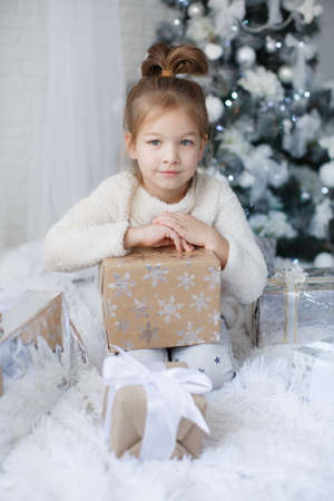 Happy child, 5 year old girl in Christmas holidays near a beautiful green Christmas tree, decorated with white toys and snowflakes, sits next to beautifulの写真素材