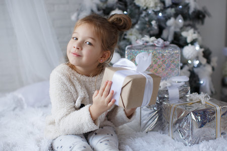 Happy child, 5 year old girl in Christmas holidays near a beautiful green Christmas tree, decorated with white toys and snowflakes, sits next to beautifulの写真素材