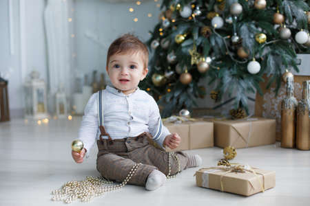 Little boy with huge brown eyes, blond short hair holding a toy for Christmas tree gold color, wearing a light brown trousers and white striped shirt, sitting on the floor next to a white Christmas tree and boxes with giftsの写真素材