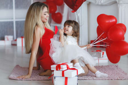 Young beautiful mother and her little daughter, dressed in a beautiful, festive and elegant dress, preparing gifts and red balloons in the form of heart for a beautiful winter holiday-Valentine's day.The concept of love in a happy familyの写真素材