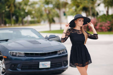 Young lady, brunette with long hair, beautiful young woman, wearing earrings in the ears is black summer dress sleeveless, head wears a big black hat, on the hand bracelet black, one travels through the resort area of ??America in a chic black convertibleのeditorial素材