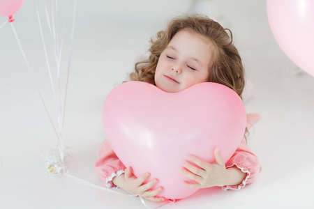 A little girl of 6 years with long curly hair, dressed in a pink dress and white tights, a beautiful smile, sits alone in a large bright room with lots of pink balloons in the shape of a heart. Valentine's Day and party celebration.の写真素材