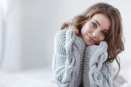 Cute young girl in a gray knitted sweater is relaxing in a white bedroom.の写真素材