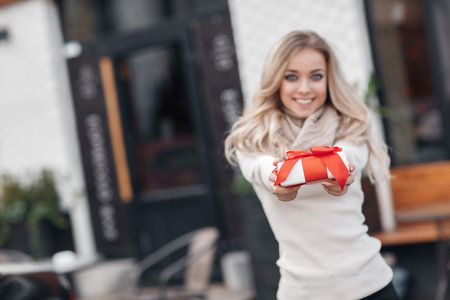 Pretty girl with long blonde hair, standing alone in the open air, holding a Christmas gift. Christmas woman with gift box.Cute young blonde in a white winter sweater with a red gift box, Christmas, New Year, Valentine's Day giftの写真素材