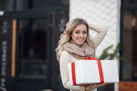 Pretty girl with long blonde hair, standing alone in the open air, holding a Christmas gift. Christmas woman with gift box.Cute young blonde in a white winter sweater with a red gift box, Christmas, New Year, Valentine's Day giftの写真素材