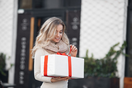 Pretty girl with long blonde hair, standing alone in the open air, holding a Christmas gift. Christmas woman with gift box.Cute young blonde in a white winter sweater with a red gift box, Christmas, New Year, Valentine's Day giftの写真素材