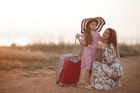 A beautiful young mother with long blond hair, a sweet smile, dressed in a light long dress, crouched on a country road next to her little daughter in the big straw hat from the sun, the girl holding a red suitcase on wheels, a travel conceptの写真素材
