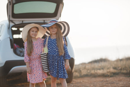 Two little girls standing on the shore of the ocean near the open trunk of the white car, loaded with stuffの写真素材