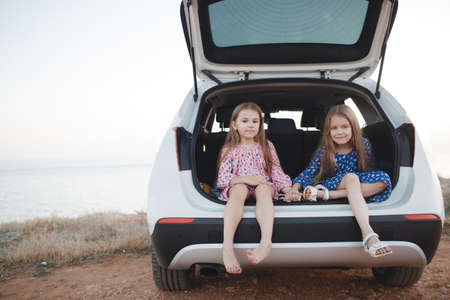 Two little girls sitting in the open trunk of the white car that is on the ocean, outdoors in the summer, things loaded, the sisters dressed in pink and blue dress with long sleeves, both girls long straight hair, travel conceptの写真素材