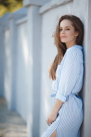 Portrait of young beautiful smiling woman, brunette with long straight hair and brown eyes, long black eyelashes dressed in a light blue dress in a white stripe, posing outdoors in summer in a green parkの写真素材