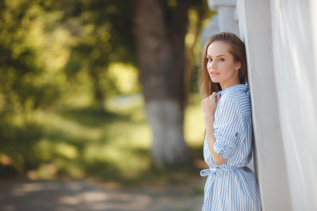 Portrait of young beautiful smiling woman, brunette with long straight hair and brown eyes, long black eyelashes dressed in a light blue dress in a white stripe, posing outdoors in summer in a green parkの写真素材