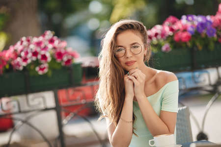 Portrait of beautiful smiling young woman with long hair, brunette, sitting at table looking in camera. Woman with cup of coffee, tea outside on summer day.の写真素材
