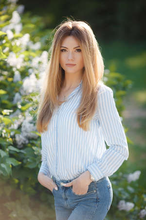 A slender, tall, beautiful woman, model appearance, long straight red hair, green eyes, dressed in a light striped shirt, blue jeans, posing in the summer outdoors, walking alone in a green park near flowering bushesの写真素材