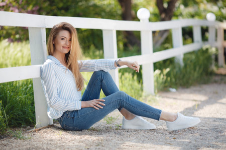 Slender, tall, beautiful woman, model appearance, long straight red hair, green eyes, wearing jewelry, dressed in a light striped shirt, blue jeans, posing in the summer outdoors, sitting alone near a white wooden fence in a green parkの写真素材