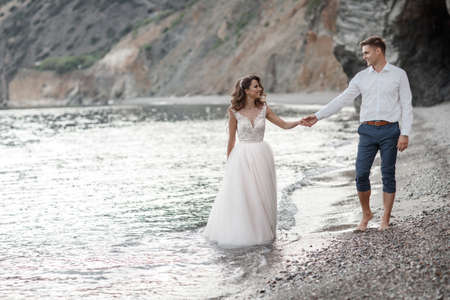 A beautiful bride and groom on their wedding day walk along a rocky shore near the sea.Young Couple On The Beach, Honeymoon, Happy Smiling Man And Woman Walking Seaside Sea Ocean Holiday Travel.Happy Wedding Day.の写真素材