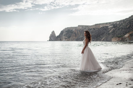 Beautiful bride stands on a cliff above the sea in a glamorous white wedding dress view of veil.Romantic beautiful bride in a white dress posing on the background sea. Happy bride in luxury dress posing on the beach.Happy Wedding Day.の写真素材