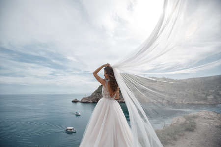 Beautiful bride stands on a cliff above the sea in a glamorous white wedding dress view of veil.Romantic beautiful bride in a white dress posing on the background sea. Happy bride in luxury dress posing on the beach.の写真素材