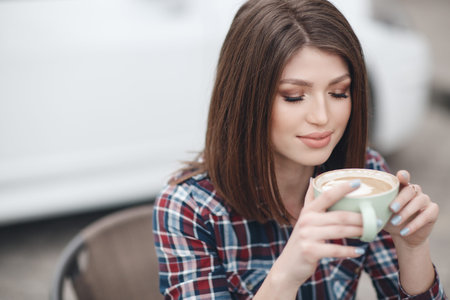 Portrait of beautiful smiling young woman with long hair, brunette, sitting at table looking in camera. Woman with cup of coffee, tea outside on summer day.Morning coffee in a summer cafeの写真素材