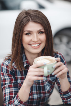 Portrait of beautiful smiling young woman with long hair, brunette, sitting at table looking in camera. Woman with cup of coffee, tea outside on summer day.Morning coffee in a summer cafeの写真素材