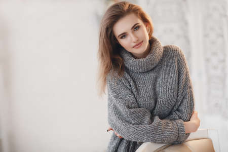 Beautiful sexy girl in an elegant gray sweater. Close-up, portrait of a beautiful woman, model, indoors. An elegant woman sits alone in a room. Attractive lady in knitted sweater posing alone, sitting in armchair at home in the roomの写真素材