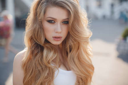 A young beautiful woman with long, wavy hair and green eyes, beautiful makeup, pink plump lips, sweet smile, blonde, spending time outdoors in the summer in the city, dressed in a white jersey, posing for a day in the sunlightの写真素材