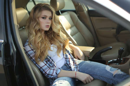 Beautiful young woman with long, thick, wavy hair, beautiful makeup with long black eyelashes, plump pink lips, wearing a light plaid shirt poses sitting on the backseat of the car hairstyle and perfect makeupの写真素材