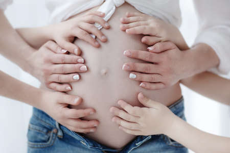 The third baby will soon be born. The arms of the young woman support the big belly of the pregnant mother. Child's hands and mother's palms lie on the belly of a pregnant womanの写真素材