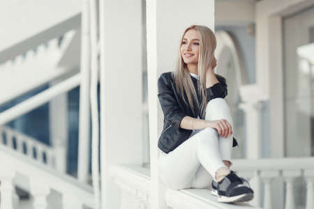 A young beautiful woman with long hair and brown eyes, beautiful makeup, pink plump lips, sweet smile, blonde, spending her time outdoors in the summer in the city, dressed in a white jersey, a black jacket and white jeans, posing on a sunny dayの写真素材