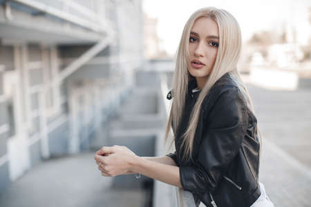 A young beautiful woman with long hair and brown eyes, beautiful makeup, pink plump lips, sweet smile, blonde, spending her time outdoors in the summer in the city, dressed in a white jersey, a black jacket and white jeans, posing on a sunny dayの写真素材