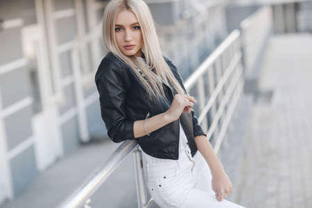 A young beautiful woman with long hair and brown eyes, beautiful makeup, pink plump lips, sweet smile, blonde, spending her time outdoors in the summer in the city, dressed in a white jersey, a black jacket and white jeans, posing on a sunny dayの写真素材