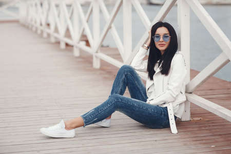 Portrait of a young, sexy, tanned beautiful girl standing on a white pier near the sea station. Beautiful woman, brunette with long hair in a white top and blue glasses.A beautiful, young woman on a sea dock waiting for her loverの写真素材