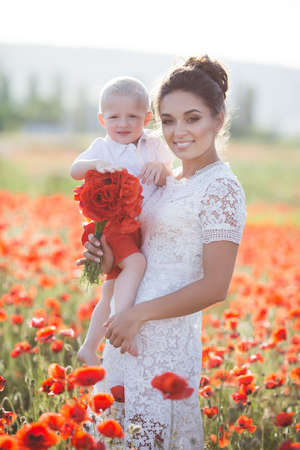A beautiful young mother in a white dress with a young son in her arms enjoying the beauty of the blooming red flowers, walking through a huge meadow on a sunny summer day.Summer scene of a happy mother and son in the field of red poppiesの写真素材