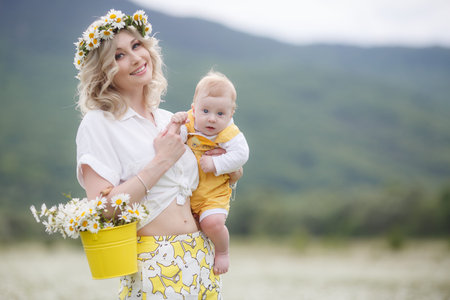 A newborn son is standing outdoors in the highlands among a blooming chamomile field. Blue sky and white meadows of blooming daisiesの写真素材