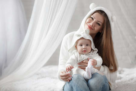 Little girl playing with her daughter at home. Attractive Caucasian woman in white top lying on her belly. Happy child playing with his mother on the white bed.の写真素材