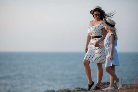 A cheerful child and her mother are enjoying the sunny morning near the water. Portrait of a beautiful brunette mother and her daughter in beautiful dresses on a rocky shore with the sea in the background. Happy family spends vacation at the resort.の写真素材