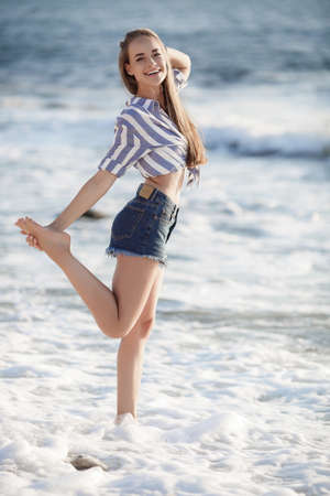 Beautiful brunette sitting on the rocks.Sexy brunette girl posing alone on the beach in the evening.Young pretty hot sexy attractive girl relaxing in in a striped shirt and shorts on stones with blue sea and sky on background.Summer Vacation Concept.の写真素材