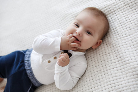 Adorable baby boy in white sunny bedroom plays alone lying on the bed.