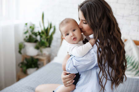 Mother and child on a white bed. Mom and baby boy in shorts playing in sunny bedroom. Parent and little kid relaxing at home.Family having fun together. Bedding and textile for infant nursery. Happy lfamily. Mother playing with her baby in the bedroomの写真素材