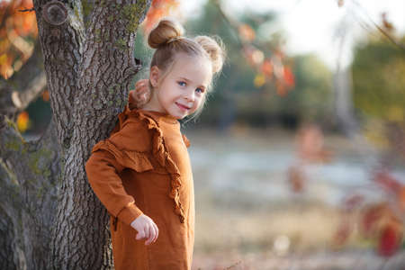 Cute girl poses for the photographer in a beautiful autumn park at sunset, walking alone. Little fashionista. Little girl happy smiling with autumn leaves. Girls autumn style.Little girl excited about autumn season.Autumn warm season pleasant moments.の写真素材