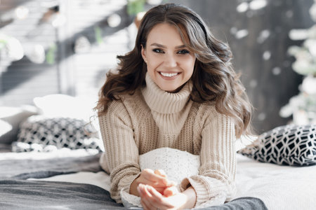 christmas and new year concept - young beautiful woman sitting in decorated living room with gift box, Christmas tree and garland lights.Portrait of beautiful young woman against christmas tree with christmas decorations.の写真素材